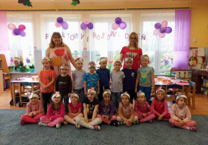 Zdjęcie grupowe – dzieci i panie w ubraniach w kolorach różowym i fioletowym.